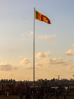 A tall flagpole with a colorful flag waves prominently against a backdrop of a partly cloudy sky. Below the flagpole is a park-like area with numerous people gathered, some playing and others walking around. In the distance, there's a bouncy castle and palm trees, contributing to a festive atmosphere. The soft golden light suggests the time is around sunset.