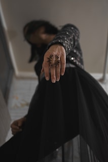 A close-up view of a person extending their hand forward, adorned with a large, ornate ring resembling a starburst. The person is dressed in a dark outfit featuring glimmering embellishments, adding a touch of sophistication. The background is a subdued indoor environment.