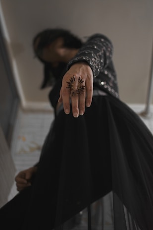A close-up view of a person extending their hand forward, adorned with a large, ornate ring resembling a starburst. The person is dressed in a dark outfit featuring glimmering embellishments, adding a touch of sophistication. The background is a subdued indoor environment.