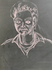 A chalk drawing depicting a man with stylized facial features including a mustache and voluminous hair. The figure is drawn on a dark surface with light detailing that highlights the contours and expressions.