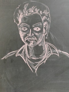 A chalk drawing depicting a man with stylized facial features including a mustache and voluminous hair. The figure is drawn on a dark surface with light detailing that highlights the contours and expressions.