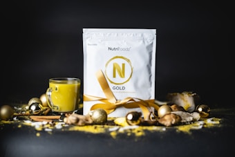 A packet labeled 'Nutrifoodz Gold' stands prominently in the center, adorned with a golden ribbon. Surrounding the packet are various festive, gold-colored ornaments and a glass mug filled with a yellow beverage. The background is dark, creating a contrast that highlights the brightness of the gold accents and the yellow drink. Cinnamon sticks and ginger pieces are scattered around, adding a cozy and warm touch to the scene.