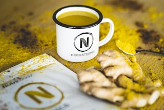 A white mug filled with a yellow liquid is placed on a surface sprinkled with yellow powder, possibly turmeric. The mug has a large 'N' logo and the hashtag #CreatingBetterHumans printed on it. Next to the mug, there is a packet with a similar logo and some pieces of ginger root. A spoon with yellow powder rests beside the ginger.