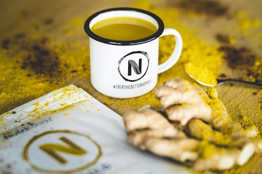 A white mug filled with a yellow liquid is placed on a surface sprinkled with yellow powder, possibly turmeric. The mug has a large 'N' logo and the hashtag #CreatingBetterHumans printed on it. Next to the mug, there is a packet with a similar logo and some pieces of ginger root. A spoon with yellow powder rests beside the ginger.