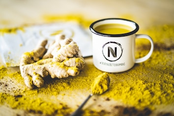 A white enamel mug filled with a yellow beverage sits on a surface covered in turmeric powder. Next to the mug, there is a piece of fresh ginger root and a spoon with turmeric powder. The mug features a logo with the letter 'N' and the hashtag '#CREATINGBETTERHUMANS'.