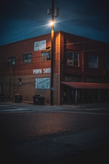 A dimly lit street corner features a brick building with signs indicating a pawn shop. The streetlight atop a wooden pole casts an orange glow over the scene, illuminating the upper part of the building and the surrounding area. Trash bins are placed near the side of the building, and the street is composed of red bricks.
