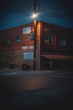A dimly lit street corner features a brick building with signs indicating a pawn shop. The streetlight atop a wooden pole casts an orange glow over the scene, illuminating the upper part of the building and the surrounding area. Trash bins are placed near the side of the building, and the street is composed of red bricks.