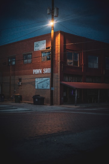 A dimly lit street corner features a brick building with signs indicating a pawn shop. The streetlight atop a wooden pole casts an orange glow over the scene, illuminating the upper part of the building and the surrounding area. Trash bins are placed near the side of the building, and the street is composed of red bricks.