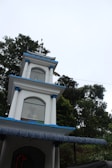 A multi-tiered white structure with blue accents and a cross on top, set against a backdrop of lush green trees under a bright sky. Each tier of the building has arched windows, with some decorative elements visible through the glass.