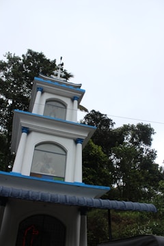 A multi-tiered white structure with blue accents and a cross on top, set against a backdrop of lush green trees under a bright sky. Each tier of the building has arched windows, with some decorative elements visible through the glass.