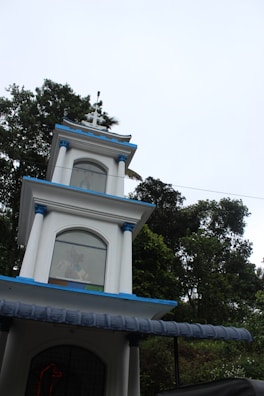 A multi-tiered white structure with blue accents and a cross on top, set against a backdrop of lush green trees under a bright sky. Each tier of the building has arched windows, with some decorative elements visible through the glass.