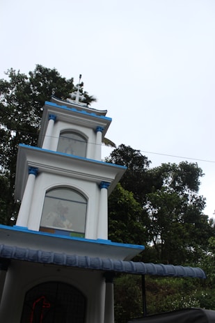A multi-tiered white structure with blue accents and a cross on top, set against a backdrop of lush green trees under a bright sky. Each tier of the building has arched windows, with some decorative elements visible through the glass.