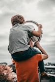 A child is sitting on top of an adult's shoulders, looking through a telescope. The adult is wearing an orange shirt, while the child wears a grey shirt. The sky is cloudy and the background is blurred, suggesting they are at an elevated viewpoint.