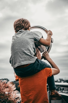 A child is sitting on top of an adult's shoulders, looking through a telescope. The adult is wearing an orange shirt, while the child wears a grey shirt. The sky is cloudy and the background is blurred, suggesting they are at an elevated viewpoint.