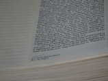 A close-up view of an open book focusing on a page with text in a two-column format. The text appears to be from a religious or spiritual scripture, with annotations or footnotes at the bottom. The left side of the page has dot matrix lines.