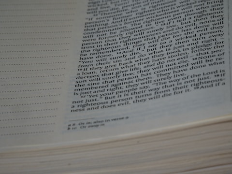 A close-up view of an open book focusing on a page with text in a two-column format. The text appears to be from a religious or spiritual scripture, with annotations or footnotes at the bottom. The left side of the page has dot matrix lines.