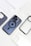 Multiple phone cases are arranged around a smartphone with a distinctive camera setup. The phone is in the center, featuring a dark case with a visible Apple logo and a circular MagSafe design. The surrounding cases are varied, including a carbon fiber pattern and several clear cases.