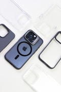 Multiple phone cases are arranged around a smartphone with a distinctive camera setup. The phone is in the center, featuring a dark case with a visible Apple logo and a circular MagSafe design. The surrounding cases are varied, including a carbon fiber pattern and several clear cases.