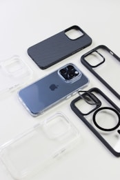 A modern smartphone with a metallic finish is placed on a white surface, surrounded by various phone cases. The phone cases come in different styles, including transparent, carbon fiber pattern, and black frames.