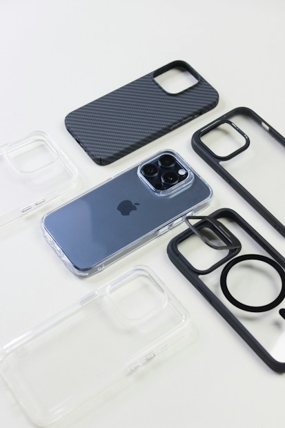 A modern smartphone with a metallic finish is placed on a white surface, surrounded by various phone cases. The phone cases come in different styles, including transparent, carbon fiber pattern, and black frames.
