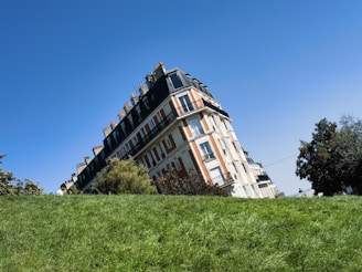 A classic Parisian-style building with a mansard roof is set against a clear, blue sky. The building features ornate detailing and multiple chimneys. In the foreground, there is a lush, green grassy area with some trees and bushes partially obscuring the building. A couple of people are visible, partially hidden by the grass.