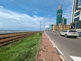 A coastal urban scene with a railway track running parallel to a road. Tall buildings with some under construction are visible, alongside a clear blue sky. The ocean is in the background, with grassy areas beside the tracks.