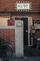 A rustic storefront with a wooden sign displaying Chinese characters is visible. The exterior features a red brick wall and a gray concrete column with the number 39 on it. A black bicycle is partially visible, leaning against the wall. The entrance is open, with some items displayed at the entrance, creating a cozy and nostalgic atmosphere.