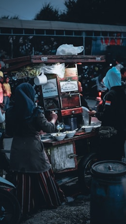 A street food vendor cart is set up with various items including bowls and cooking utensils. Two people are interacting with the cart, one appearing to serve food while the other waits. The scene is dimly lit, suggestive of an evening setting, with several people and a motorcycle visible in the background.