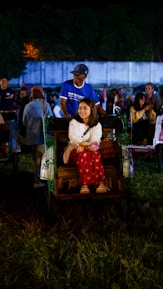 A woman is sitting comfortably in a rickshaw, while a man stands behind her, seemingly the driver. The setting appears to be a lively outdoor event at night, with other people and rickshaws visible in the background. The atmosphere seems festive and cheerful.