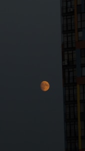 A partially illuminated moon appears in the dark night sky, positioned next to a tall building with several visible windows. The moon has an orange hue, and the building has a modern design with darkened glass panels.