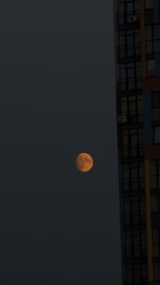 A partially illuminated moon appears in the dark night sky, positioned next to a tall building with several visible windows. The moon has an orange hue, and the building has a modern design with darkened glass panels.