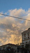 The image captures a cityscape with a dominant view of vintage-style buildings under a sky filled with fluffy, golden clouds. The lighting suggests a sunset, casting a warm glow over the scene. A strong, dark power line stretches across the sky, contrasting with the soft clouds.