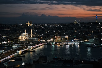 A stunning cityscape at dusk with a prominent mosque illuminated in the foreground, a bridge crossing a body of water, and a darkening sky with hints of orange and purple. The skyline is dotted with various lights, creating a lively yet serene atmosphere.