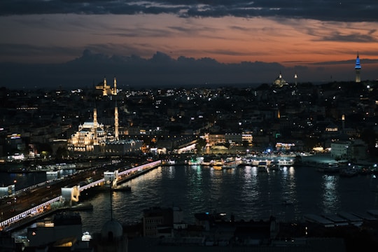 A stunning cityscape at dusk with a prominent mosque illuminated in the foreground, a bridge crossing a body of water, and a darkening sky with hints of orange and purple. The skyline is dotted with various lights, creating a lively yet serene atmosphere.