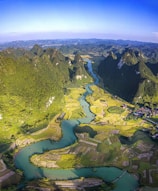 A lush green valley with a winding river flowing through terraced fields. The landscape is surrounded by towering, jagged mountains covered with dense vegetation. The fields vary in shades of green and golden yellow, indicating a mix of crops. A small village with colorful houses is nestled at the base of the mountains.