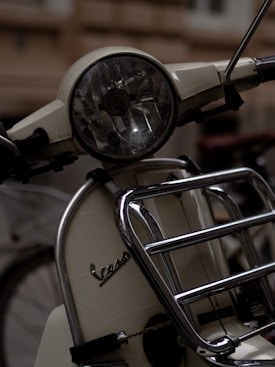 Close-up of a classic scooter's front, featuring a round headlight and chrome handlebars with a metal grill. The cream-colored body has vintage styling with a logo on the front.