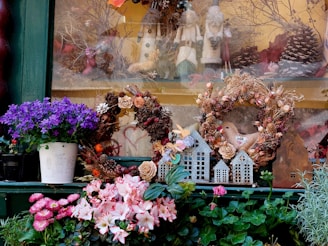 A decorative display featuring two wreaths made of dried flowers and natural materials, complemented by small decorative houses and a fabric bird. Purple and pink flowers in pots add vibrant color to the scene, while figurines and pinecones in the background provide a festive touch.