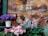 A decorative display featuring two wreaths made of dried flowers and natural materials, complemented by small decorative houses and a fabric bird. Purple and pink flowers in pots add vibrant color to the scene, while figurines and pinecones in the background provide a festive touch.