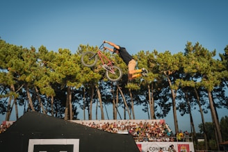A BMX rider is performing a trick in mid-air against a backdrop of tall green trees. The rider is holding the handlebars of a bicycle, with legs extended backward, showcasing a high level of skill and control. In the background, there is a crowd of spectators standing behind a barricade, watching the performance.