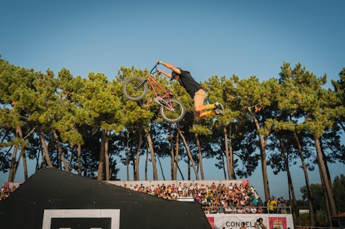 A BMX rider is performing a trick in mid-air against a backdrop of tall green trees. The rider is holding the handlebars of a bicycle, with legs extended backward, showcasing a high level of skill and control. In the background, there is a crowd of spectators standing behind a barricade, watching the performance.