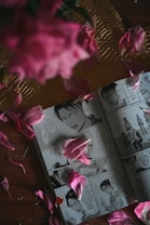 A manga book is open to a page showing detailed illustrations of characters engaging in a conversation. Pink flower petals are scattered artistically over the pages, contributing to a visually appealing arrangement. The background includes a textured, golden surface that contrasts with the wooden table beneath.