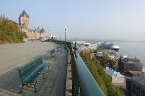 A historic hotel stands prominently to the left, with a boardwalk leading towards a gazebo adorned with a Canadian flag. The view overlooks a river, with several large cruise ships docked at the port. Green benches line the wooden walkway, and the area is surrounded by trees and buildings.