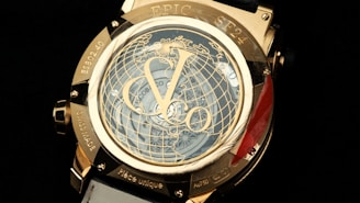 A luxury watch features intricate detailing with a prominent globe design on its face. The numerals and watch hands are elegantly integrated, surrounded by a metallic gold casing. There are engraved words and numbers on the case, and the visible strap is white.