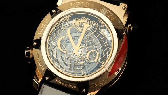 A luxury watch features intricate detailing with a prominent globe design on its face. The numerals and watch hands are elegantly integrated, surrounded by a metallic gold casing. There are engraved words and numbers on the case, and the visible strap is white.