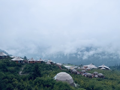 A picturesque mountain landscape with several dome-shaped structures and tents nestled among lush greenery. Mist and clouds hover low in the sky, creating a serene and tranquil atmosphere.