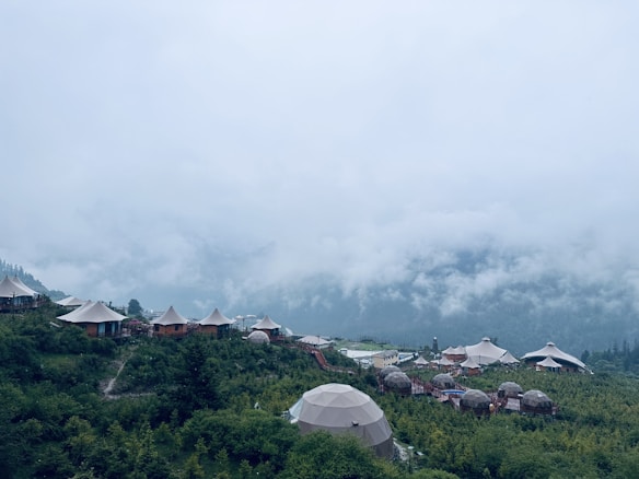 A picturesque mountain landscape with several dome-shaped structures and tents nestled among lush greenery. Mist and clouds hover low in the sky, creating a serene and tranquil atmosphere.