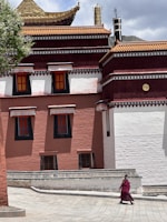 A Buddhist monk wearing traditional maroon robes walks along a spacious courtyard in front of a large, intricately designed building with red and white walls and a tiered roof adorned with gold accents. There is a tree with green leaves on the left side, providing shade.