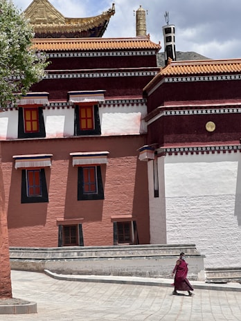 A Buddhist monk wearing traditional maroon robes walks along a spacious courtyard in front of a large, intricately designed building with red and white walls and a tiered roof adorned with gold accents. There is a tree with green leaves on the left side, providing shade.