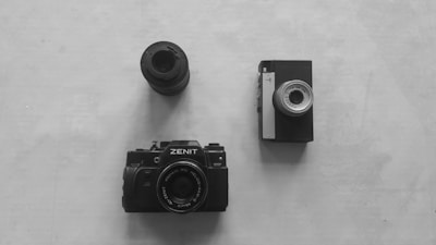 On a light surface, three vintage photographic items are arranged: a camera labeled 'Zenit' at the bottom, a lens at the top, and a retro camera on the right.