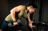 A person is engaged in a workout session, holding a dumbbell with one hand while leaning on a bench. They are wearing a green sleeveless shirt and dark shorts, with earbuds in their ears. The setting appears to be a gym, with a focused and determined expression on their face.